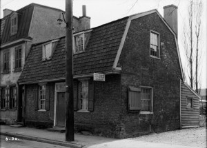 A black and white photograph of a building from the front. The image shows a side of the house, and buildings and landscaping around the building are visible.