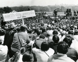 a black and white photograph of a large crowd seated in front of a stage. The stage has several stacks of amplifiers. Crew members are working on the stage. In the foreground, a man holds a sign reading "earth day every day. Don't litter."