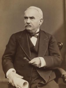 This black and white photograph shows Morton McMichael in a seated position. He holds spectacles in one hand and a rolled up newspaper in the other.