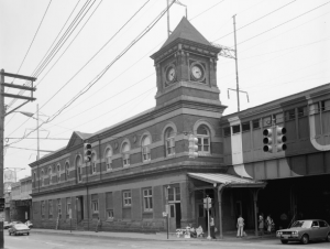 A black and white photograph of the wilmington train station showing a three story clock tower, covered railway on the second story, and entrances on the ground floor.